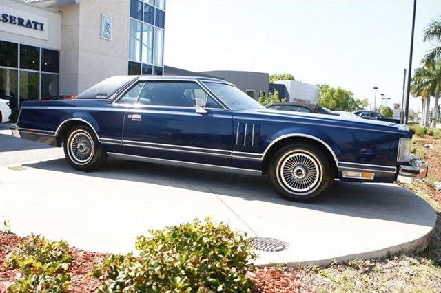 A classic navy blue vintage car parked outdoors near a building with large glass windows and palm trees.