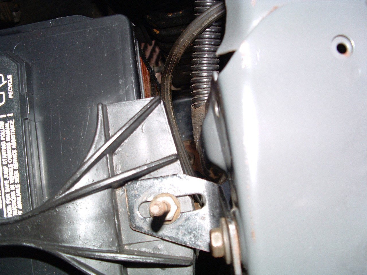 Close-up of a car battery mounted near the engine compartment with visible wires, bolts, and metal brackets.
