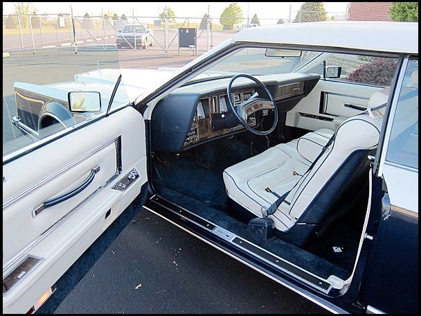 Interior of a vintage white car parked outdoors, showing the steering wheel, dashboard, front passenger seat, and door panel.