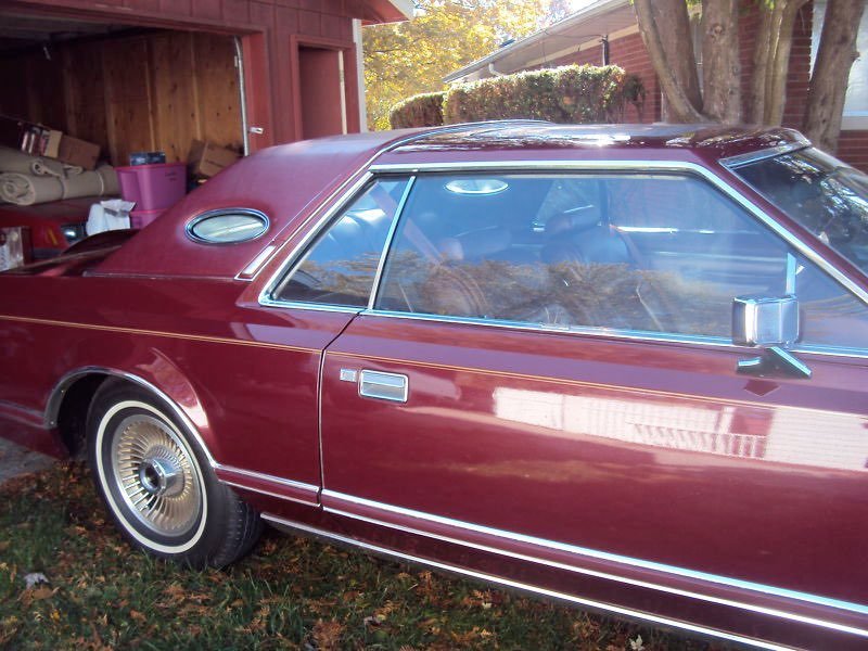 A vintage maroon car with chrome trim parked outside, next to a wooden shed and a house, with autumn trees in the background.
