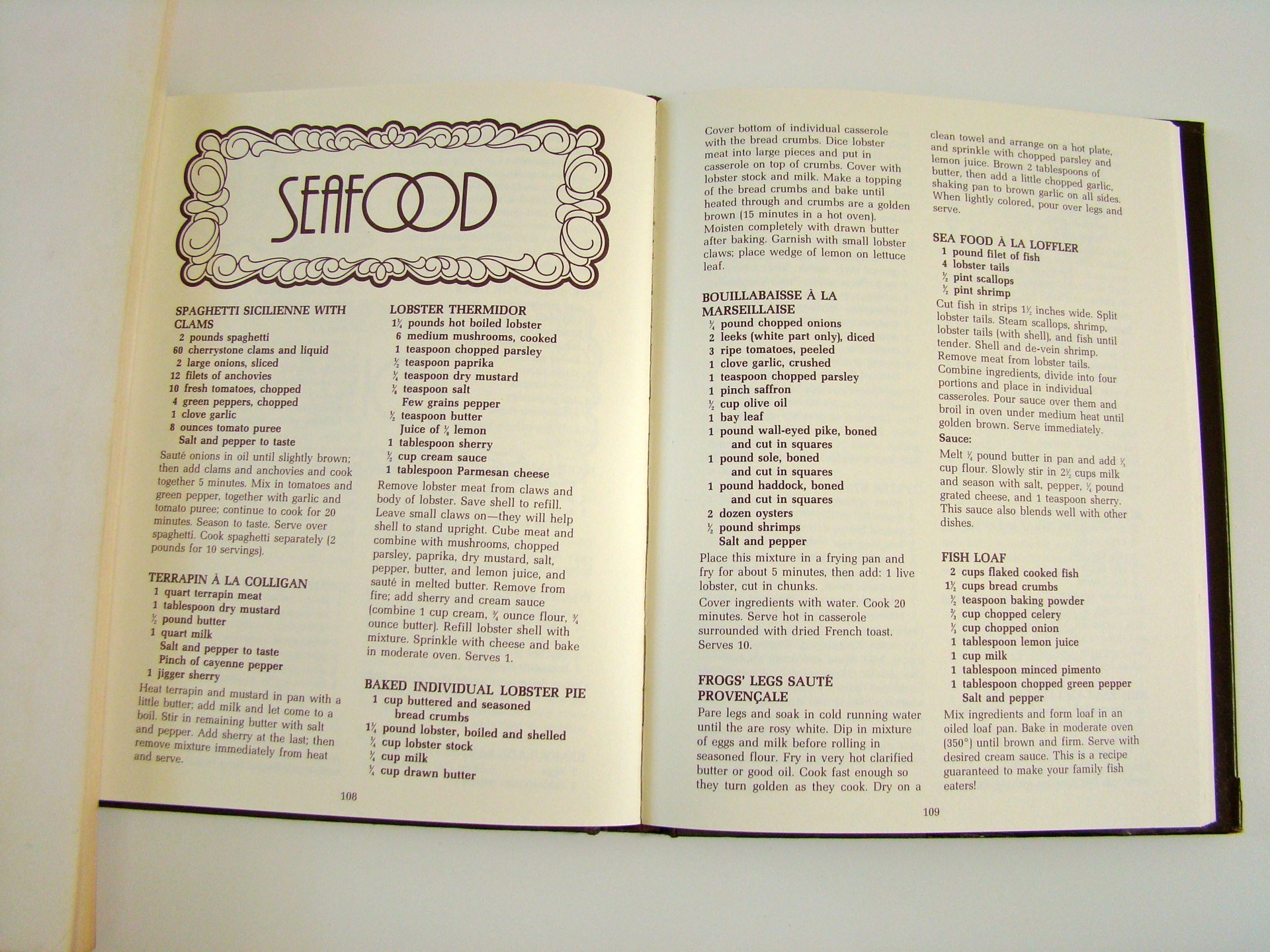 Open cookbook page titled 'Seafood' with various seafood recipes, including Spaghetti Sicilienne with Clams, Lobster Thermidor, Bouillabaisse à la Marseillaise, Fish Loaf, and Frog's Legs Sauté Provençale.
