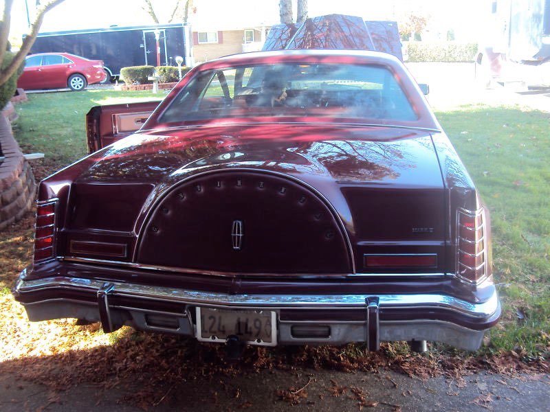 Black vintage Lincoln car parked on grass, with a spare tire cover on the trunk, and a license plate visible, surrounded by trees and other parked vehicles.