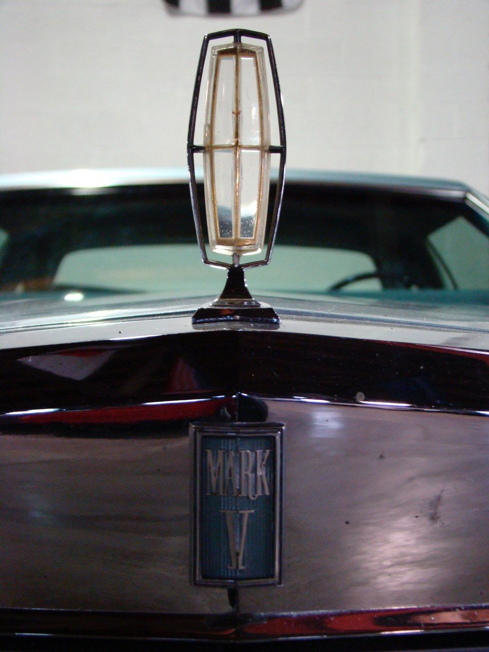Close-up of a vintage car hood ornament and badge, featuring a geometric light fixture on top and a Mark V emblem on the front.