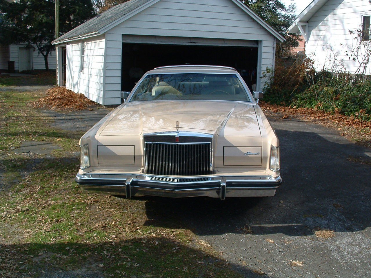 A beige vintage Lincoln Continental car parked in front of a garage with a partly open door, in front of a white house with trees and plants around.