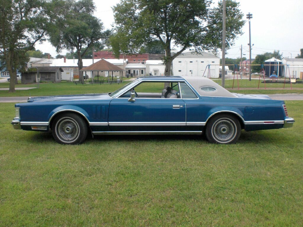 A vintage blue car with a gray vinyl roof parked on grass in a park with trees, playground equipment, and small buildings in the background.