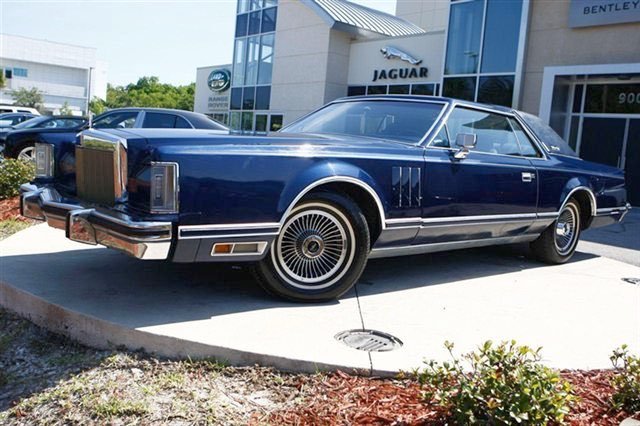 A vintage dark blue luxury car parked outside a Jaguar dealership. The car has a classic boxy design, large chrome grille, and distinctive wire-spoke hubcaps.