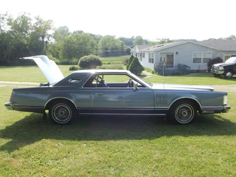 A vintage dark green car with an open hood and closed trunk parked on a grassy lawn in front of a house, with trees and other vehicles in the background.