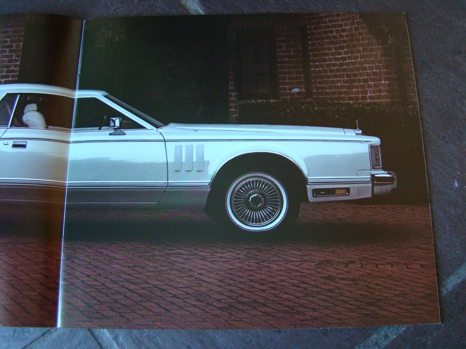 A vintage white car with a sleek design parked on a brick street against a brick wall background.