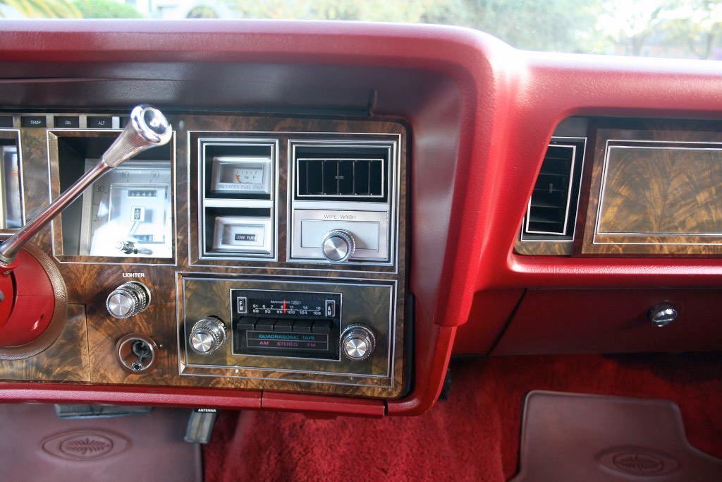 Close-up of a vintage car dashboard with wood grain paneling, red trim, various dials, switches, and a radio.