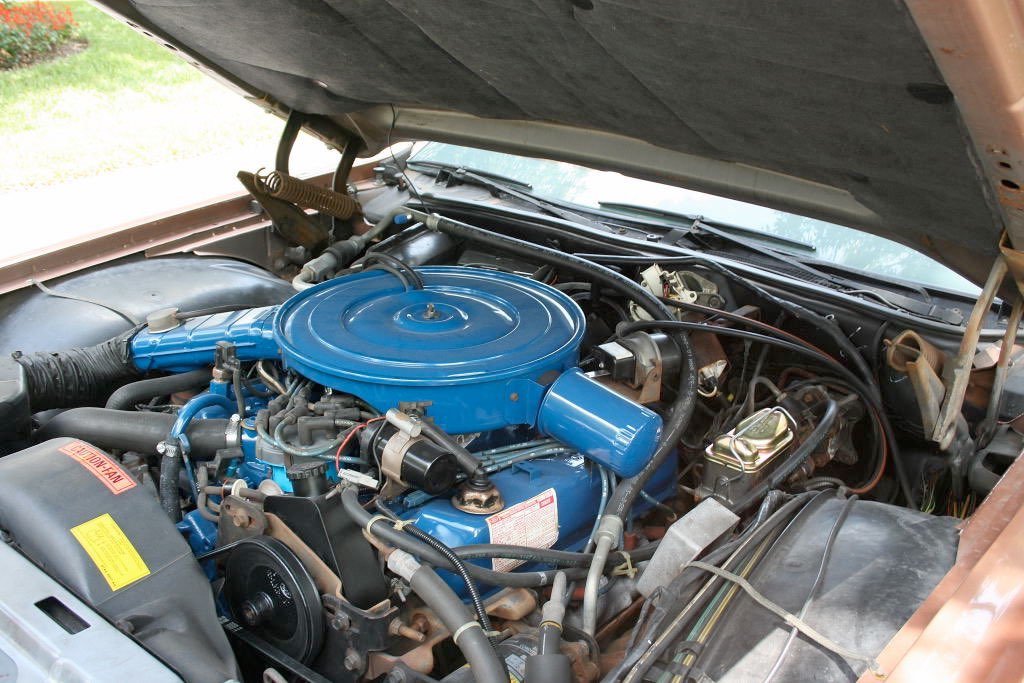 Under the hood of a vintage car showing a bright blue engine with various hoses, wires, and mechanical parts.