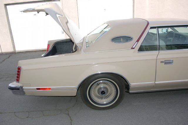 A beige vintage Lincoln Continental coupe with its rear trunk open, parked on a concrete surface near a white wall and garage door.