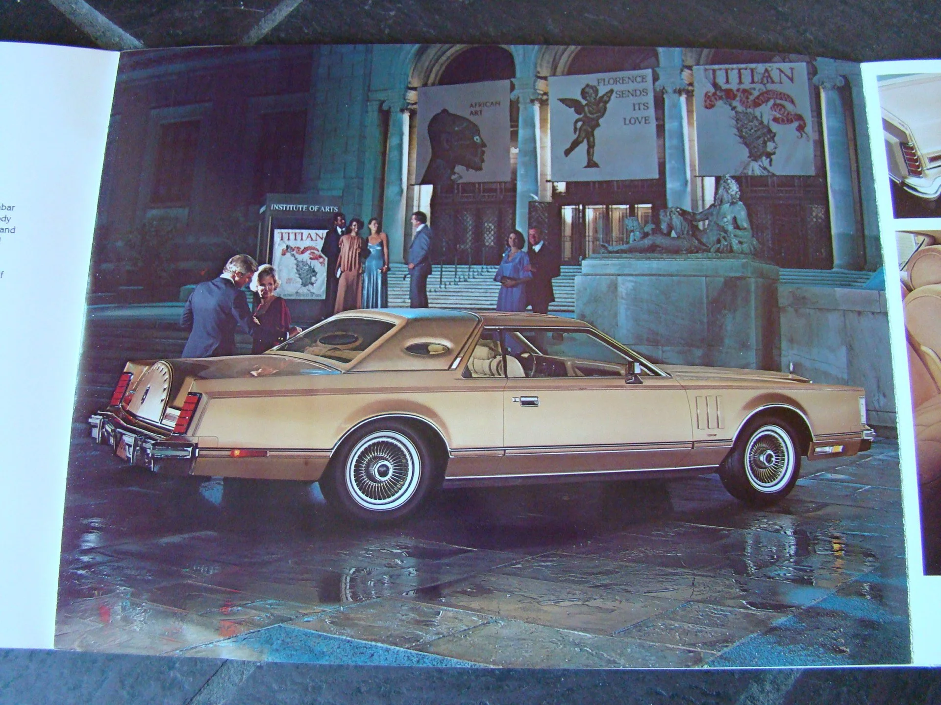 A vintage cream-colored car parked near the steps of an art institute with several people dressed in formal attire, some talking and others walking, in front of banners and sculptures outside the building.