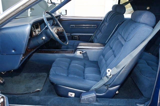 Interior of a vintage car showing the driver's seat, steering wheel, and passenger seats with blue upholstery.