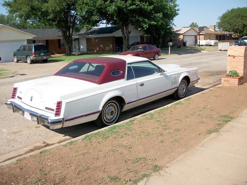 A vintage white car with a red roof parked on a residential street, with houses, trees, and other cars in the background.