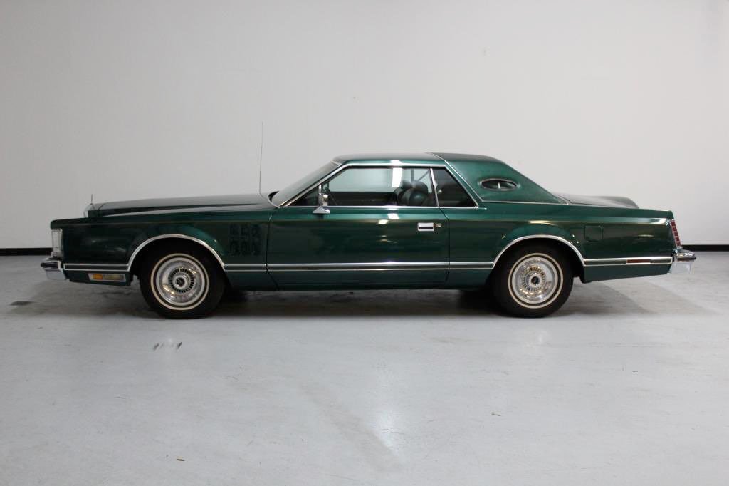 A vintage green two-door coupe car with a vinyl roof, parked indoors against a plain white wall.