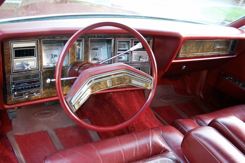 Interior of a vintage car with a red steering wheel, woodgrain dashboard, and red upholstered seats.