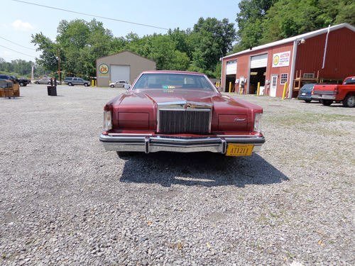 Front view of a vintage red car parked on gravel in a rural area, with buildings and trees in the background.