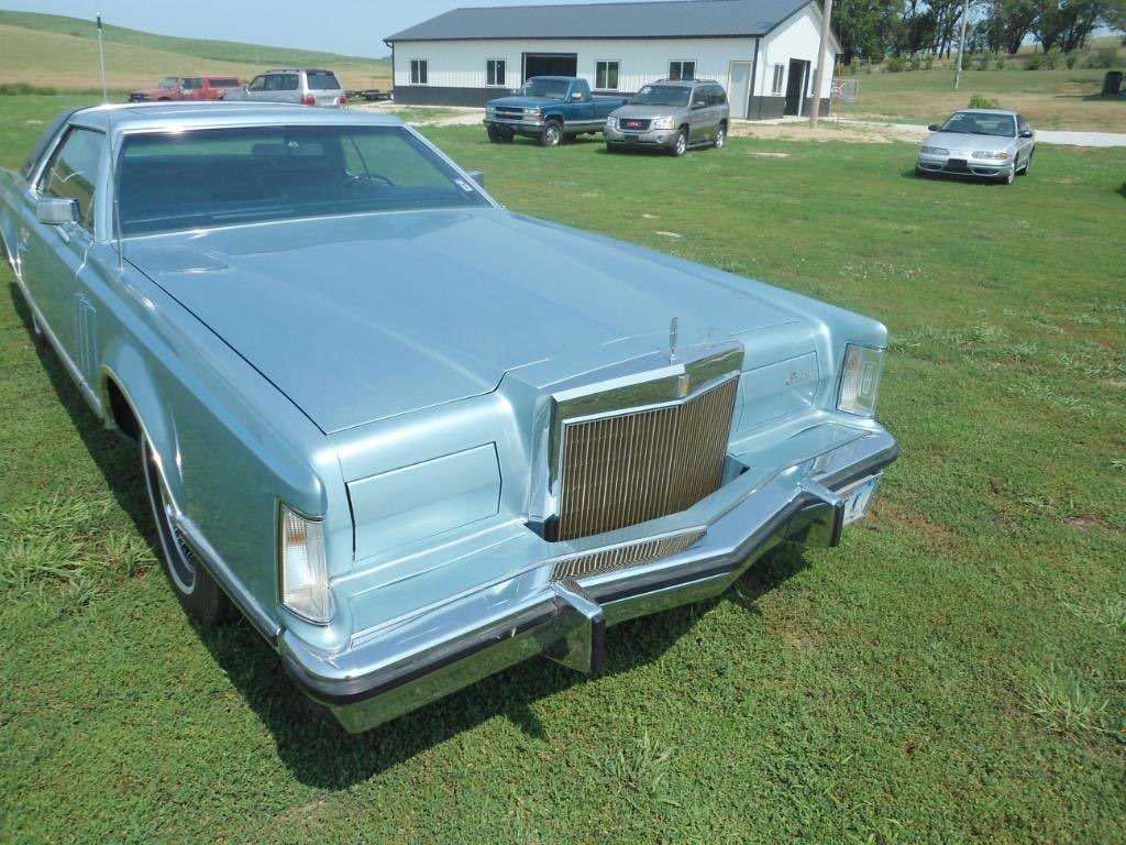 A vintage light blue classic Cadillac parked on grass with other vehicles and a white building in the background.