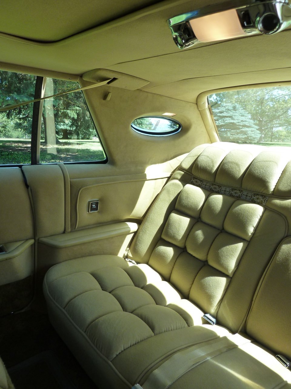 Interior of a vintage luxury car with beige plush seating, sunlit through large side and small oval rear windows, with trees visible outside.