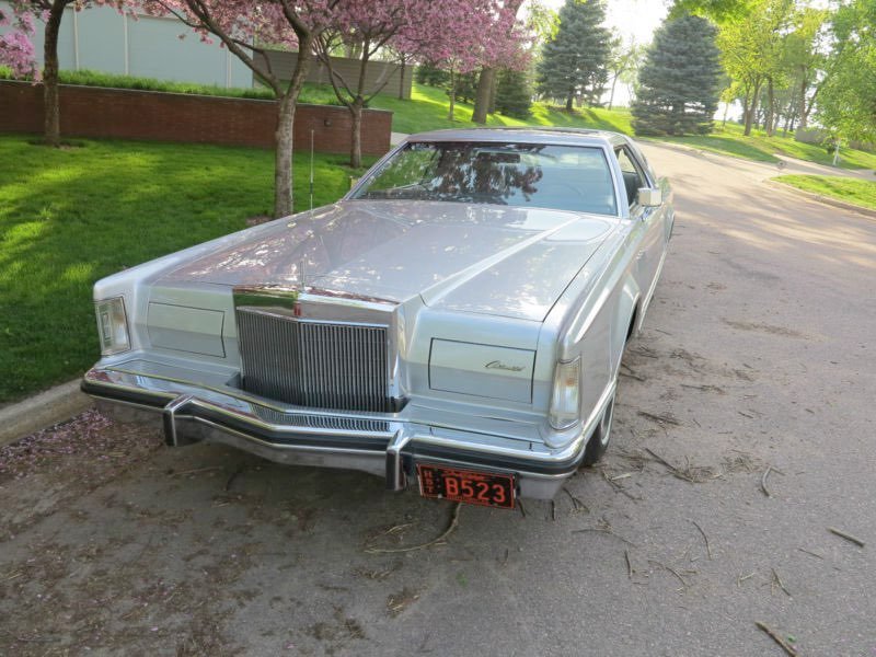 A classic silver Lincoln Continental car parked on a residential street with green grass, trees, and pink blossom trees in the background.