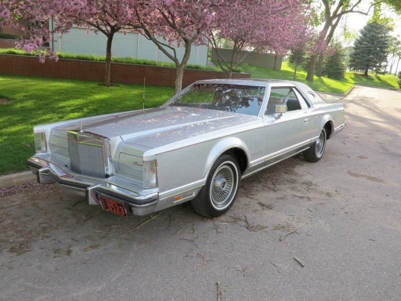 A vintage silver classic car parked on the side of a street with blooming pink trees in the background.