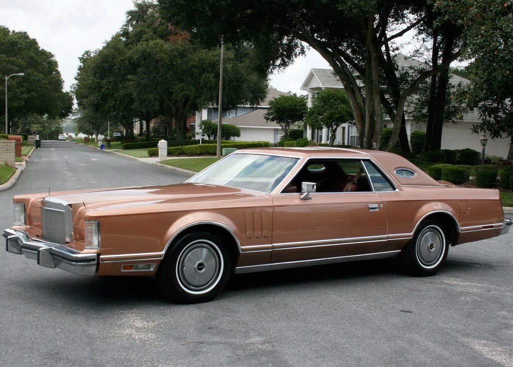 A vintage, brown, two-door luxury car with chrome accents is parked on a suburban street lined with trees and houses.