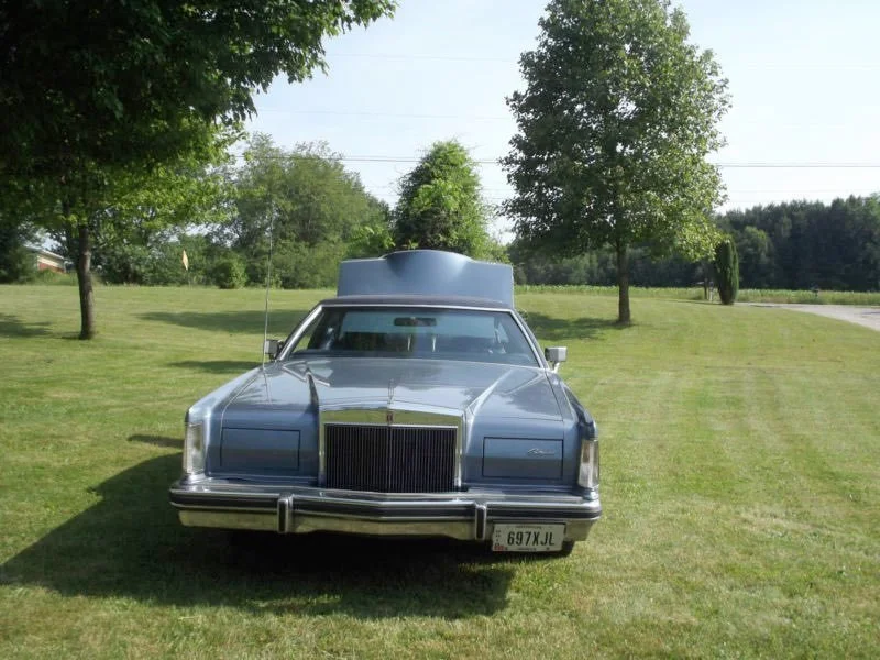 A silver vintage car with a modified body, sitting on a grassy lawn with trees in the background.