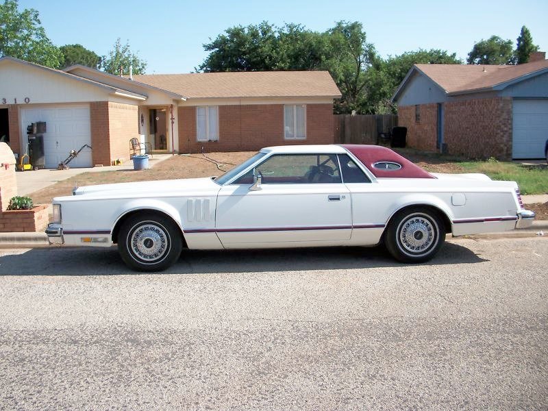 A vintage white car with a maroon roof parked on a residential street in front of a brick house with a brown roof.