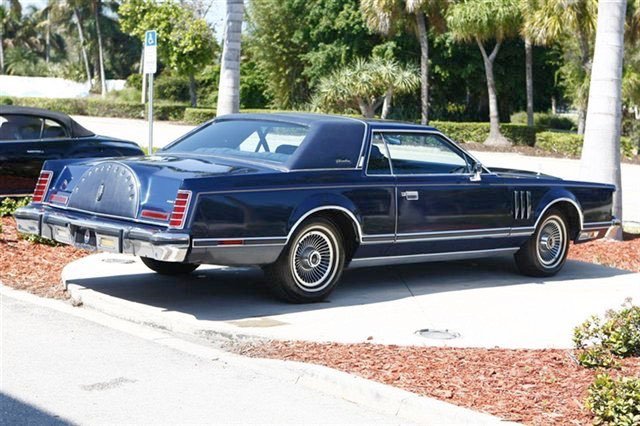 A vintage dark blue car with a canvas top parked in a parking lot with palm trees in the background.