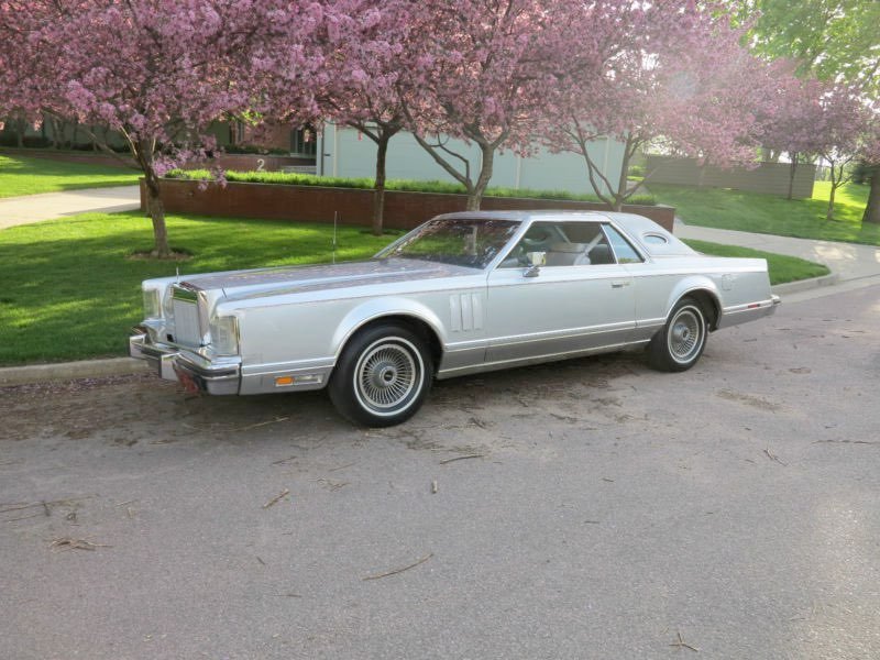 A silver vintage car parked on the side of a residential street, with pink flowering trees and green grass in the background.