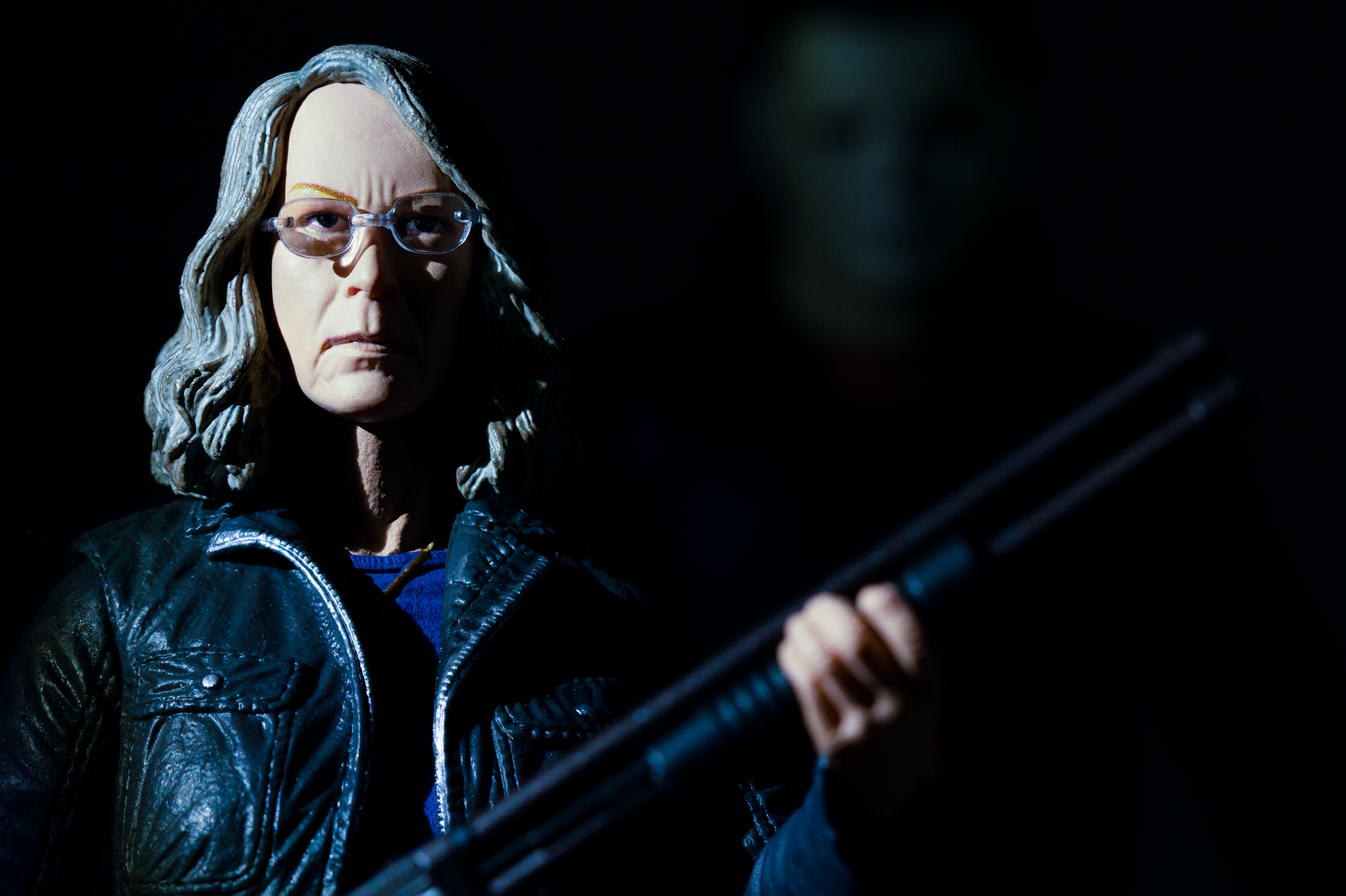 Close-up of a figure of a woman wearing glasses and a jacket, holding a shotgun in low light; behind her, a barely visible masked face emerges from the darkness.