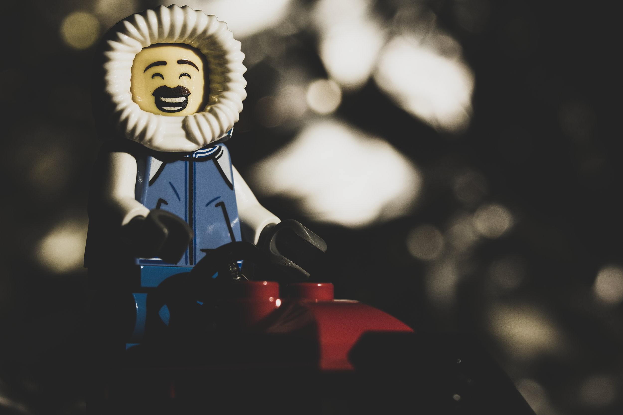 Close-up of a smiling LEGO minifigure wearing a fur-lined hood and blue jacket, seated on a red snowmobile; the background is reflective aluminum foil creating bright, abstract highlights.