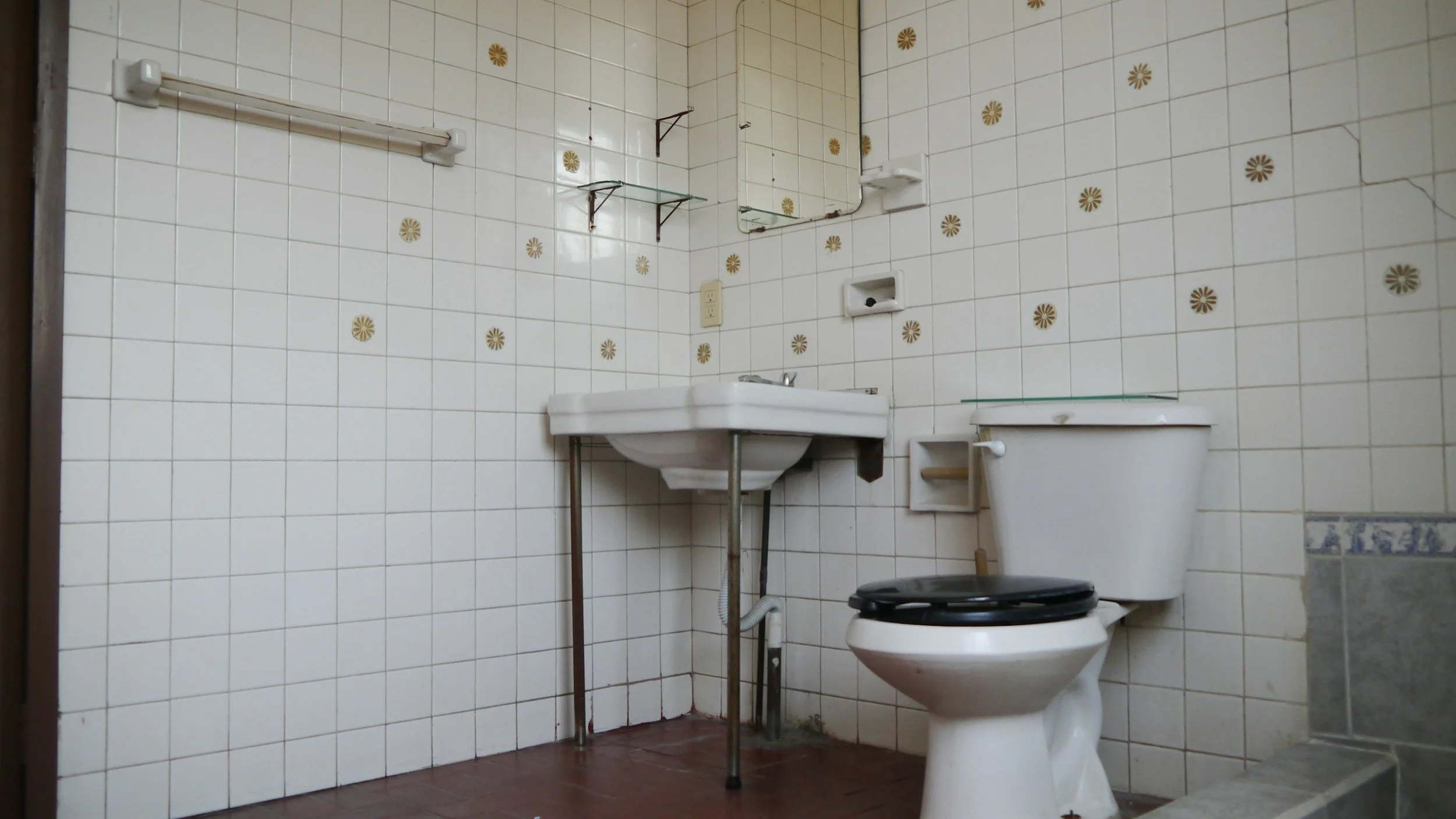 A small bathroom with white tiled walls decorated with brown floral patterns. It contains a white sink with metal support brackets, a toilet with a black seat cover, a mirror, a glass shelf, and a towel rack.