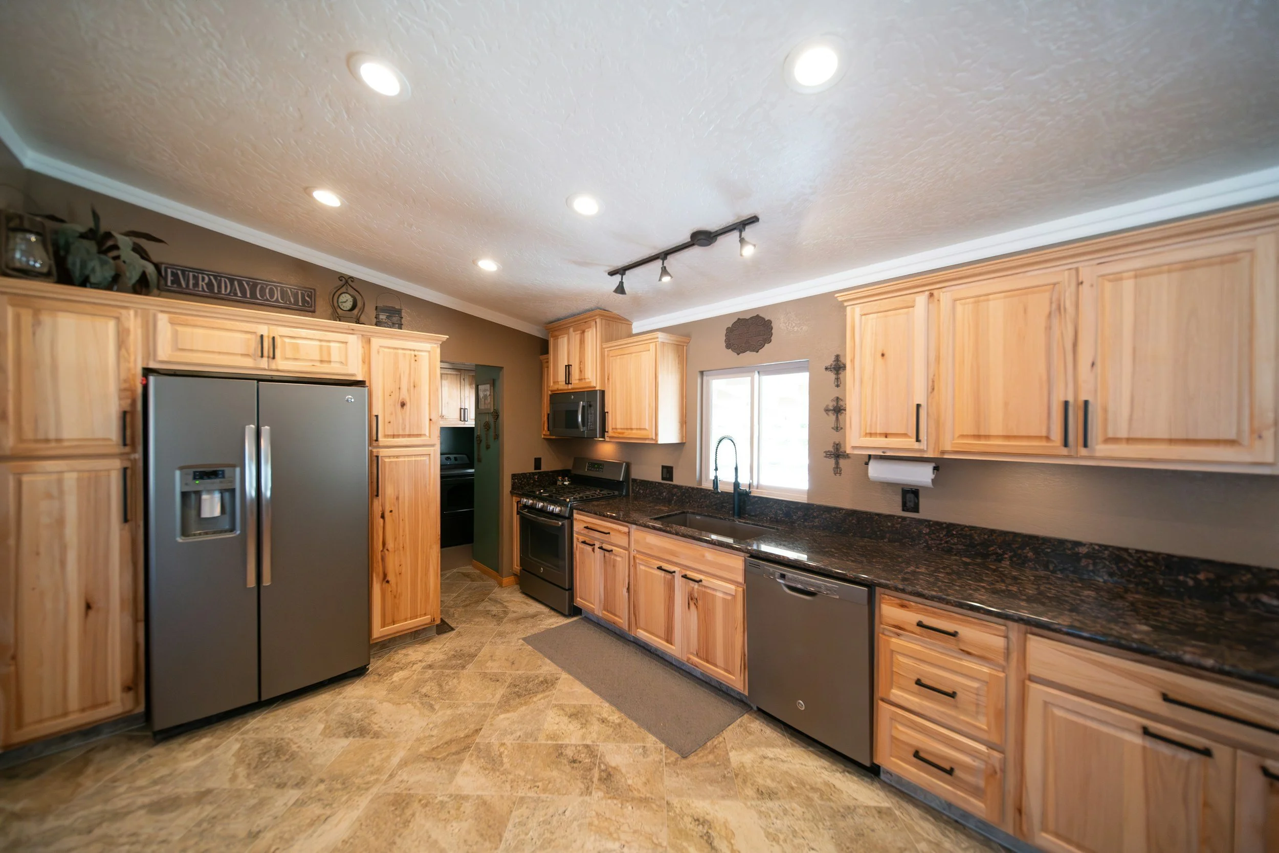 Kitchen with wooden cabinets, granite countertops, stainless steel appliances including a refrigerator and dishwasher, black stove and microwave, window above the sink, and decorative wall crosses.