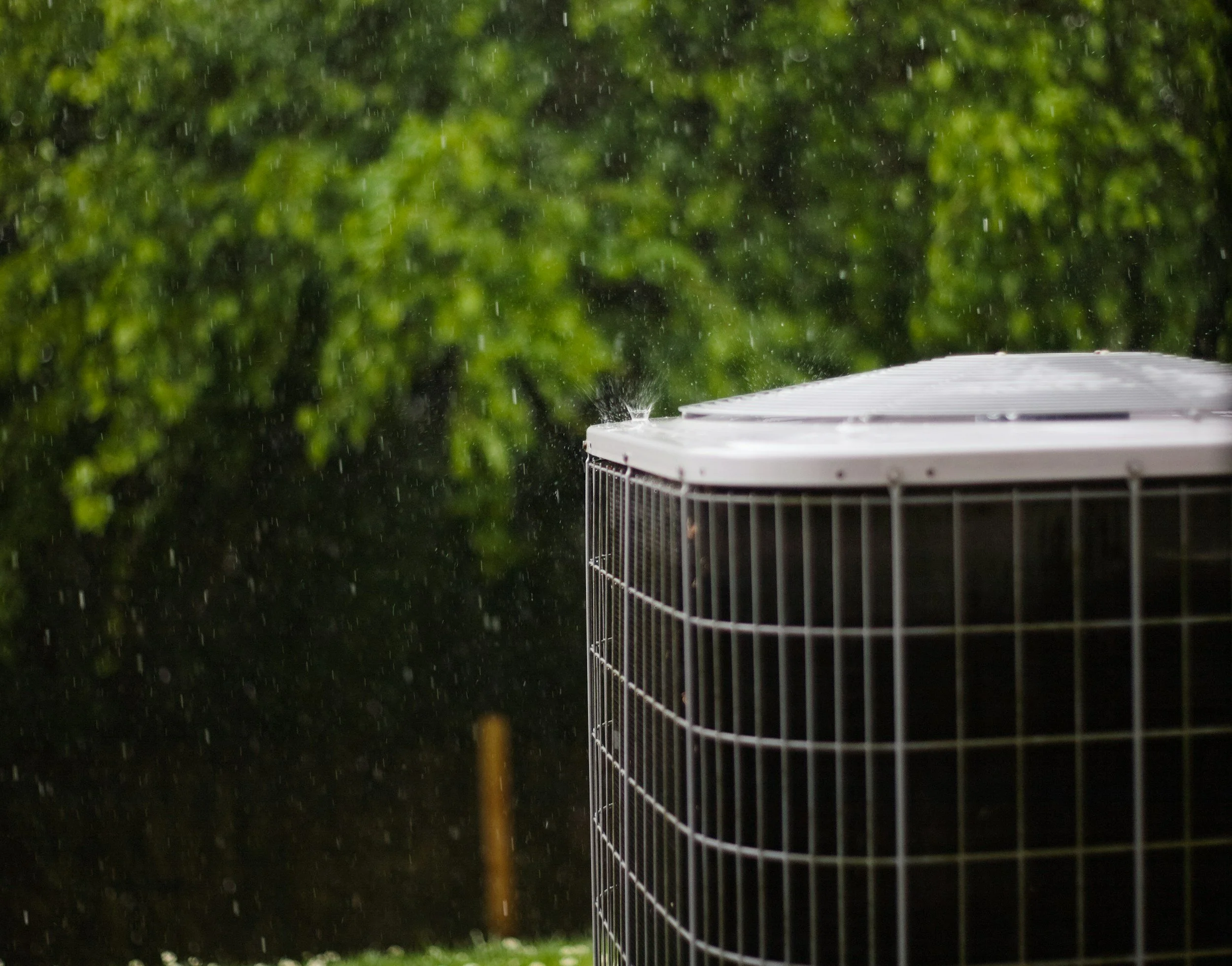 Rain falling on an outdoor air conditioning unit with a lush green background.
