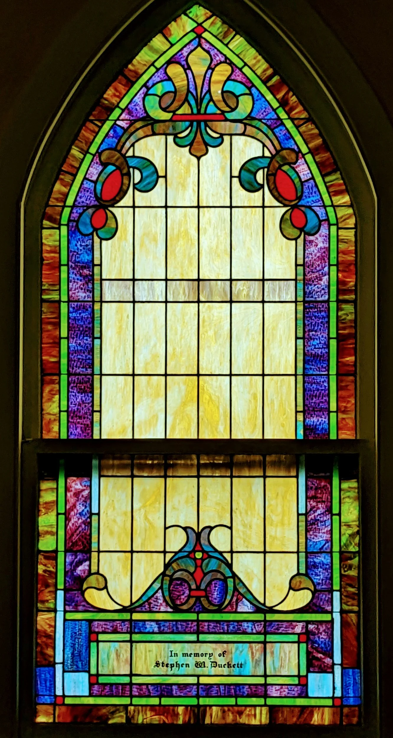 Colorful stained glass window with ornate patterns and a memorial plaque reading, 'In memory of Stephen M. Duckett'.