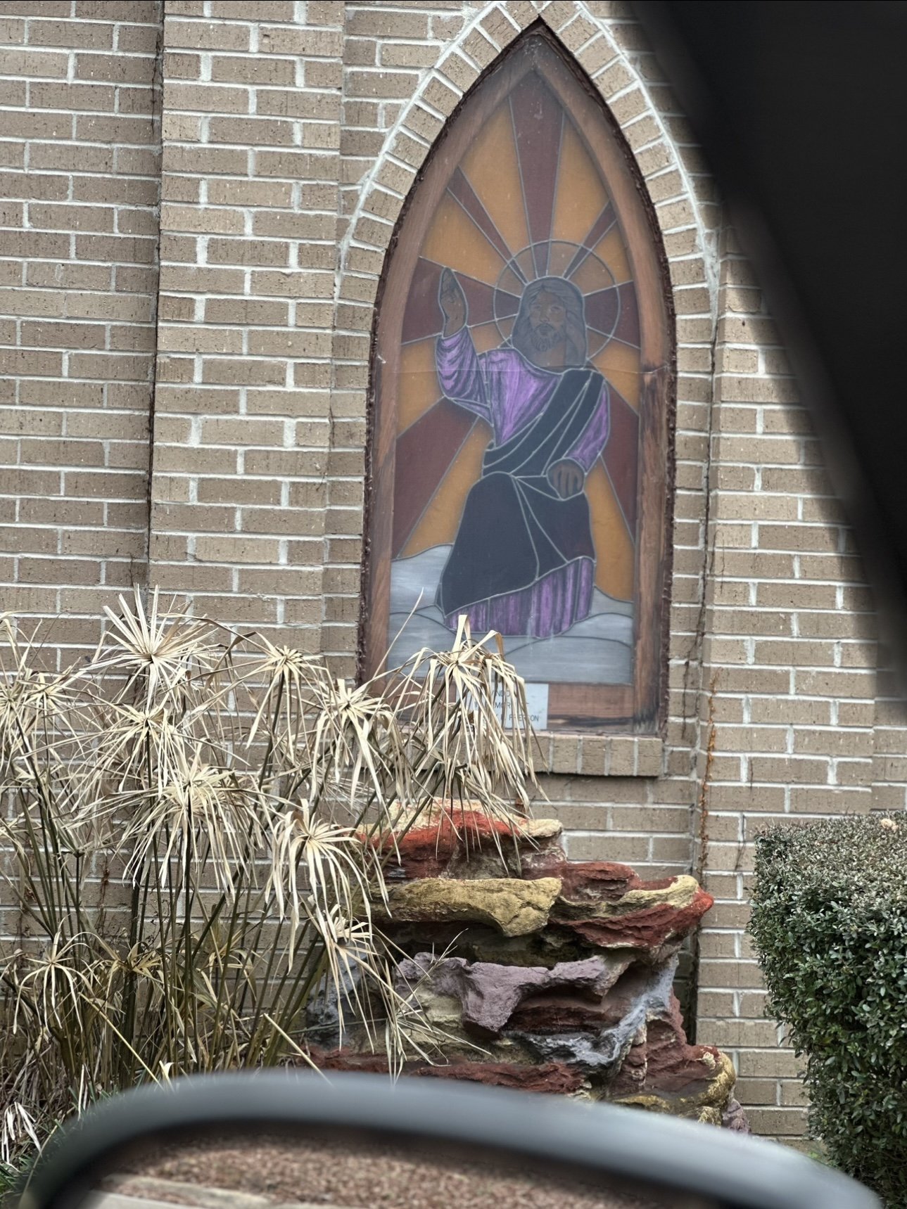 A stained glass depiction of Jesus Christ, standing on clouds with one hand raised in blessing, behind a brick church wall.