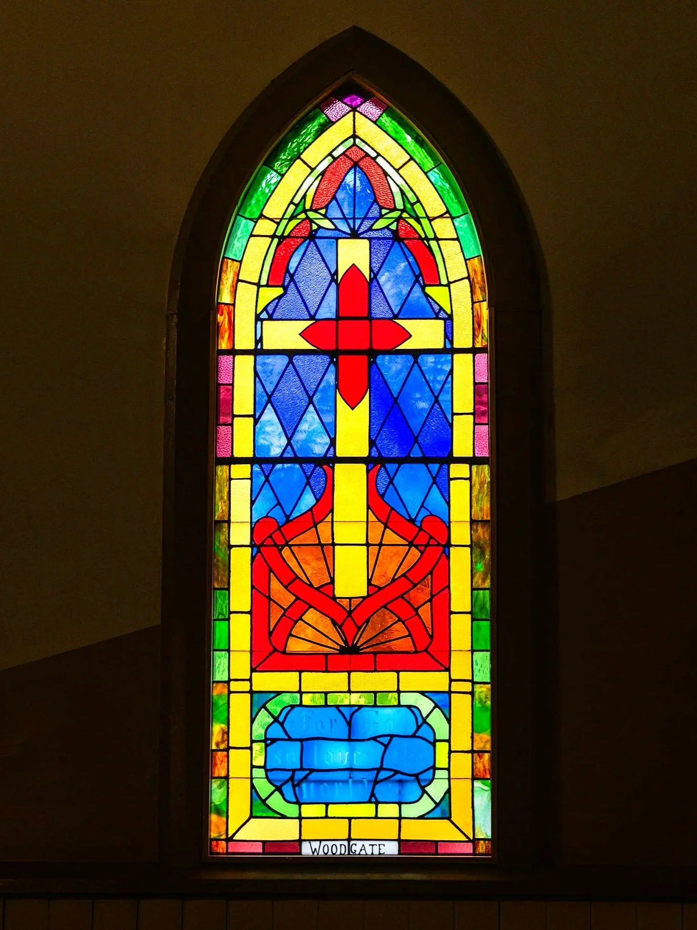 Colorful stained glass window with geometric and abstract shapes, featuring a red cross, yellow sword, red and emboldened designs, and a blue shape at the bottom, with the word 'WOODGATE' at the bottom.