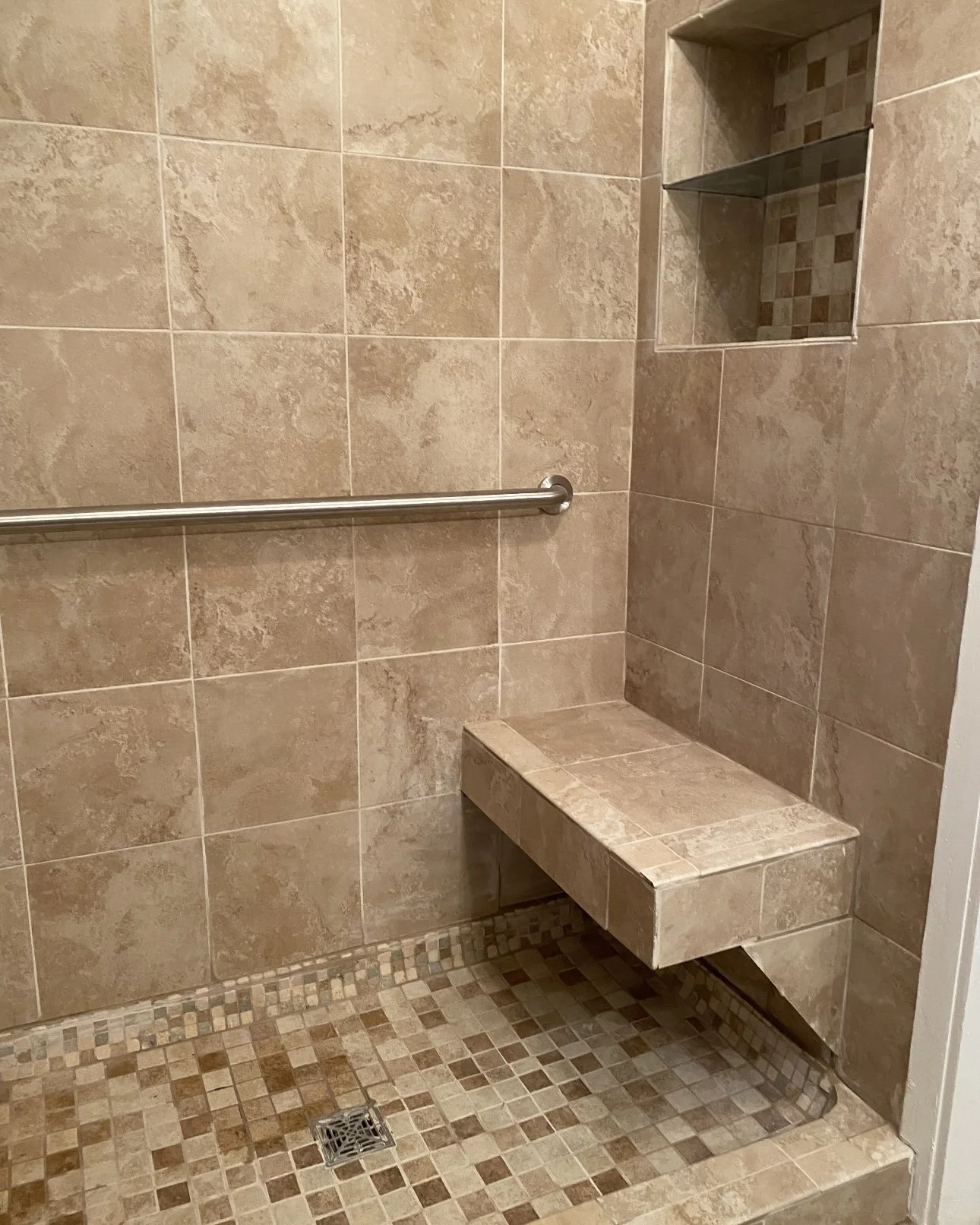 A tiled shower with beige and brown tiles, a metal grab bar, a built-in tiled bench, and a wall niche with small mosaic tiles.