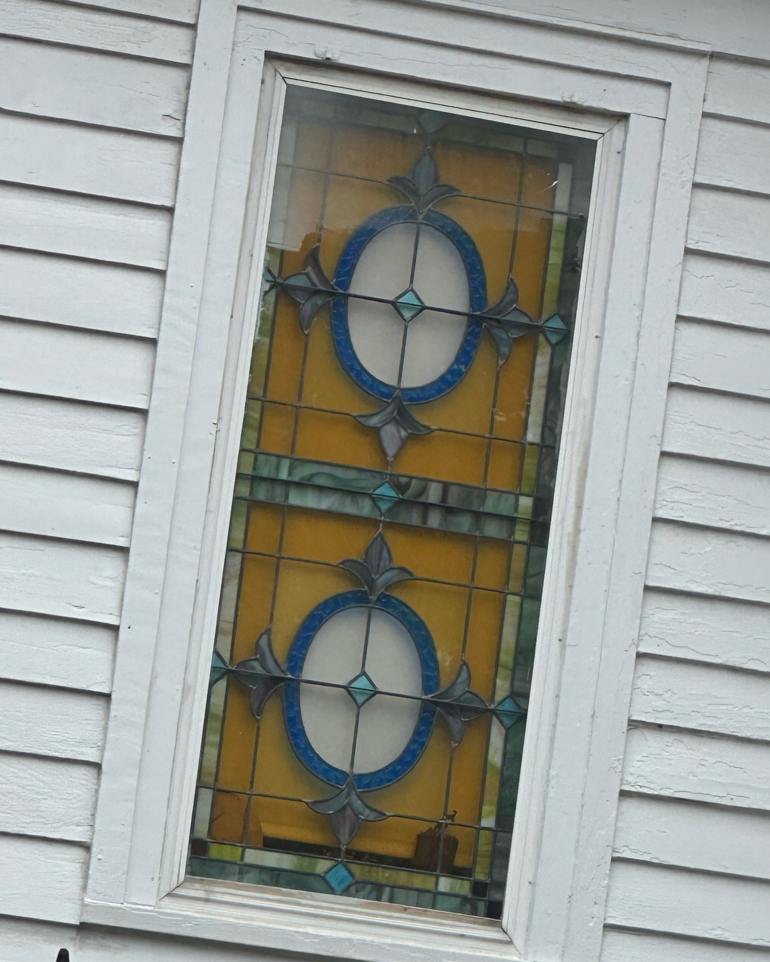 Decorative stained glass window with a yellow background and blue, gray, and clear glass accents on a white wooden wall.