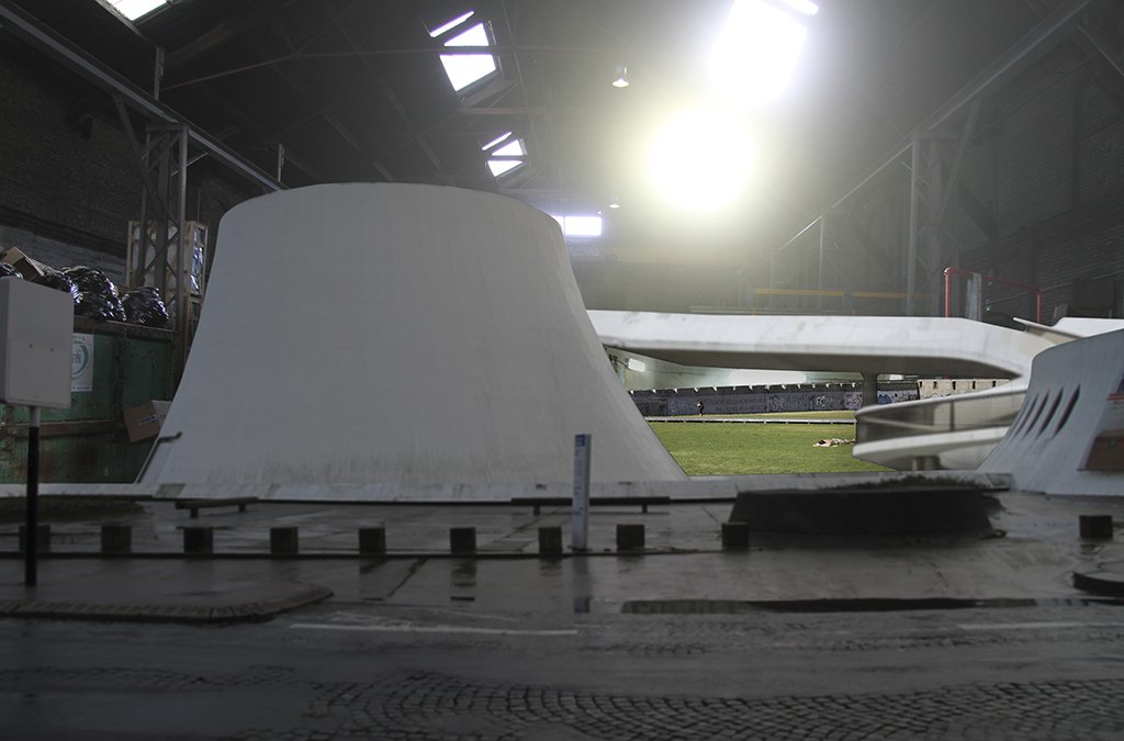 An indoor sports stadium with a section of a running track and a large, white, slanted architectural structure, possibly part of a training or event setup, with sunlight streaming in through the roof.