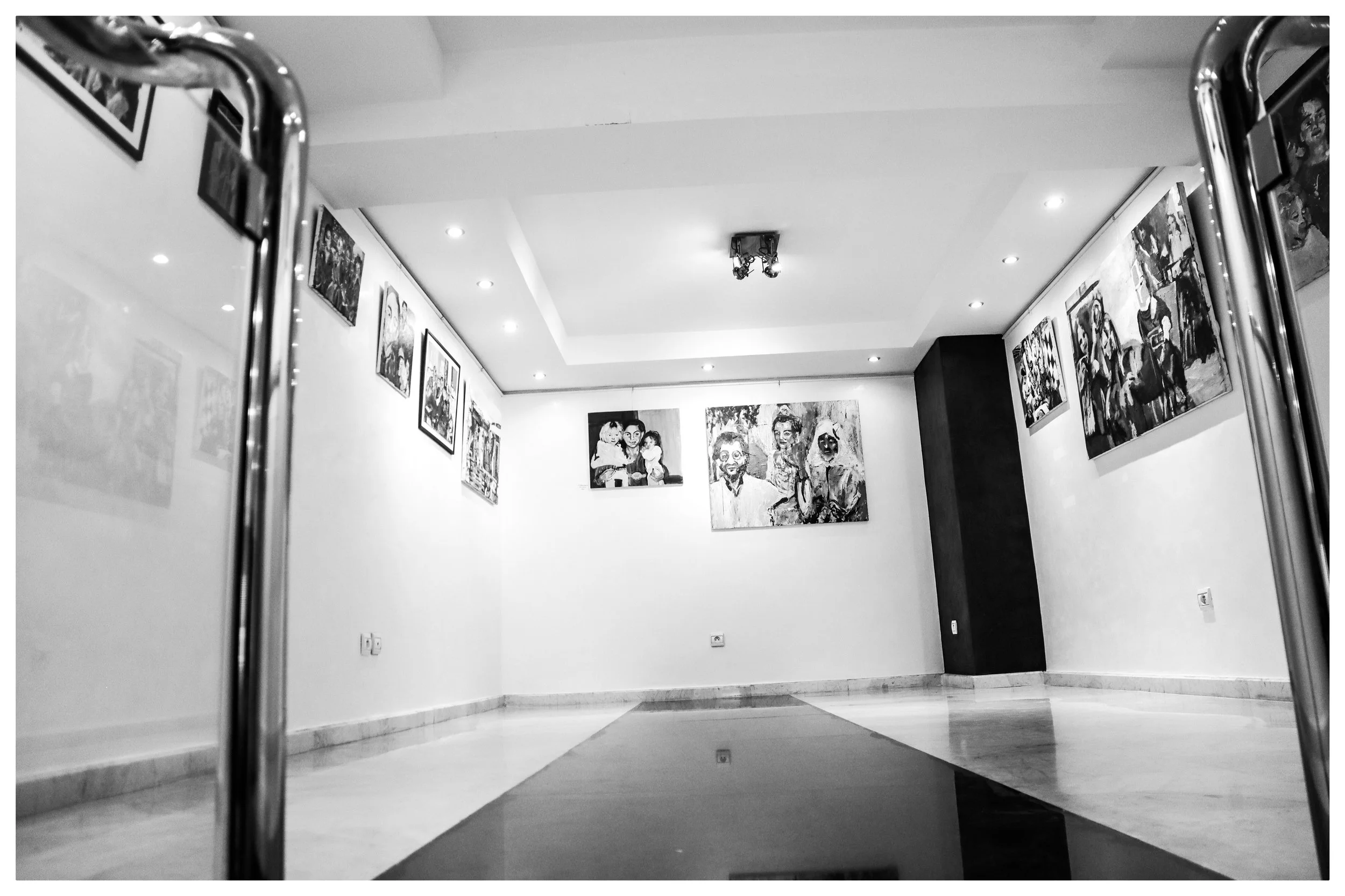 Black and white photo of an art gallery with paintings on white walls and a polished floor, viewed between the edges of a metal frame in the foreground.