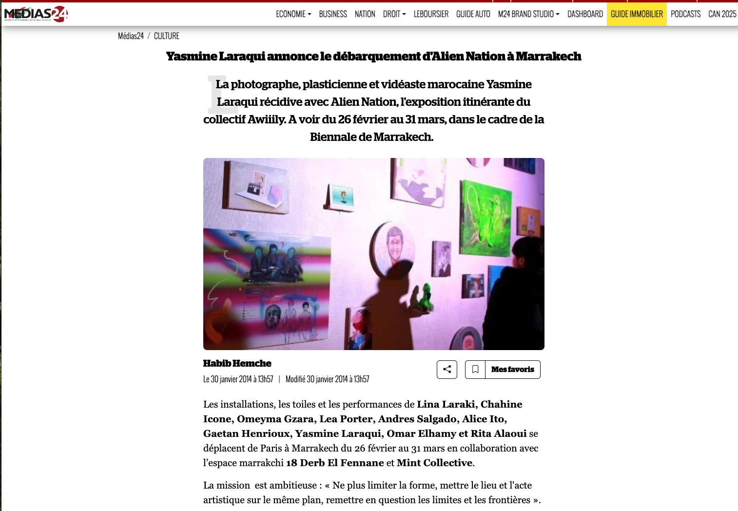 Photo of an art exhibition with various colorful artworks, including paintings and digital art, displayed on a wall. The setting appears to be an indoor gallery space with some visitors observing the art.
