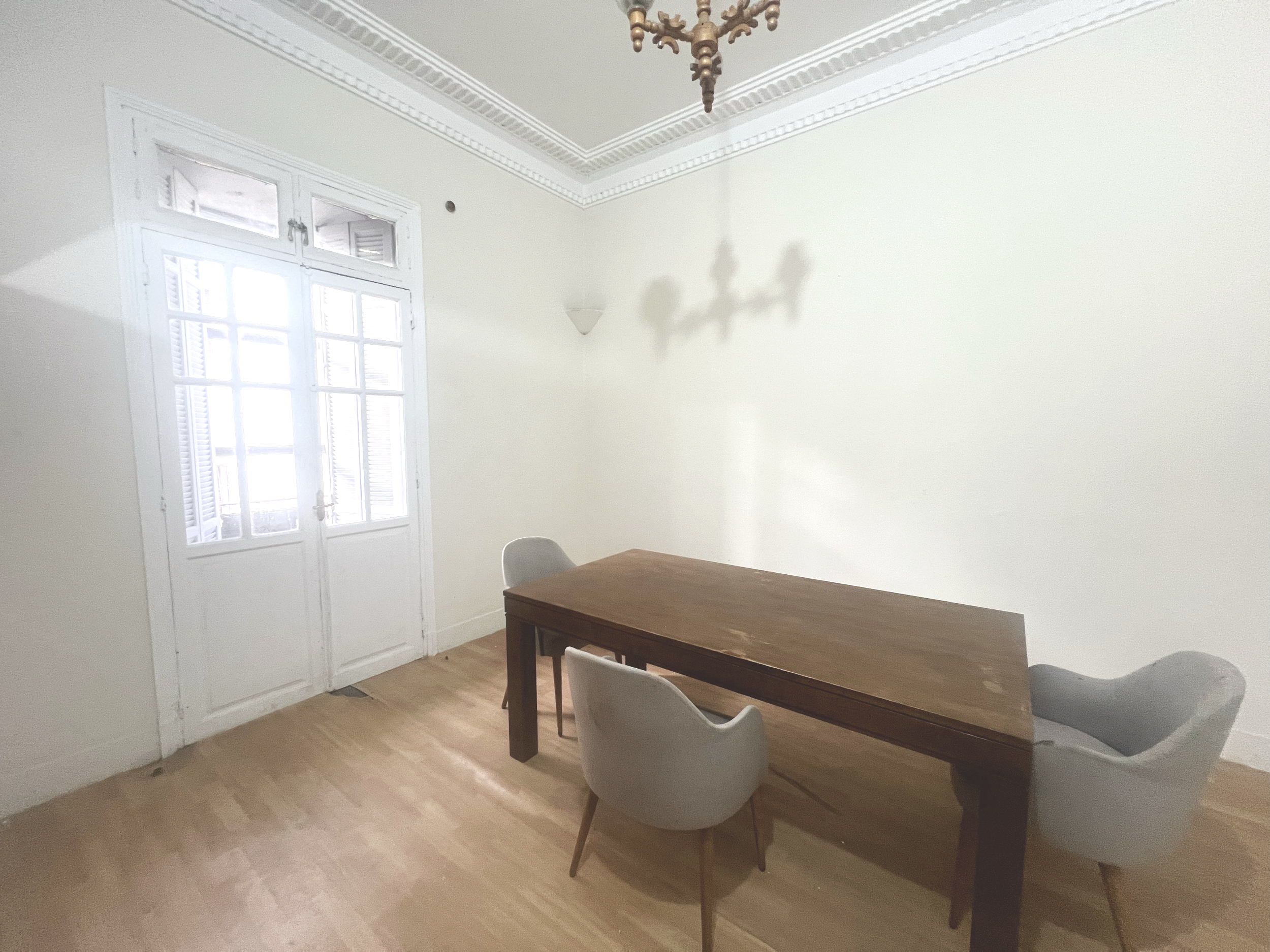 A minimalist dining room with a wooden table, three light gray upholstered chairs, a large window with white framing, wooden flooring, white walls, and a chandelier casting shadows on the wall.