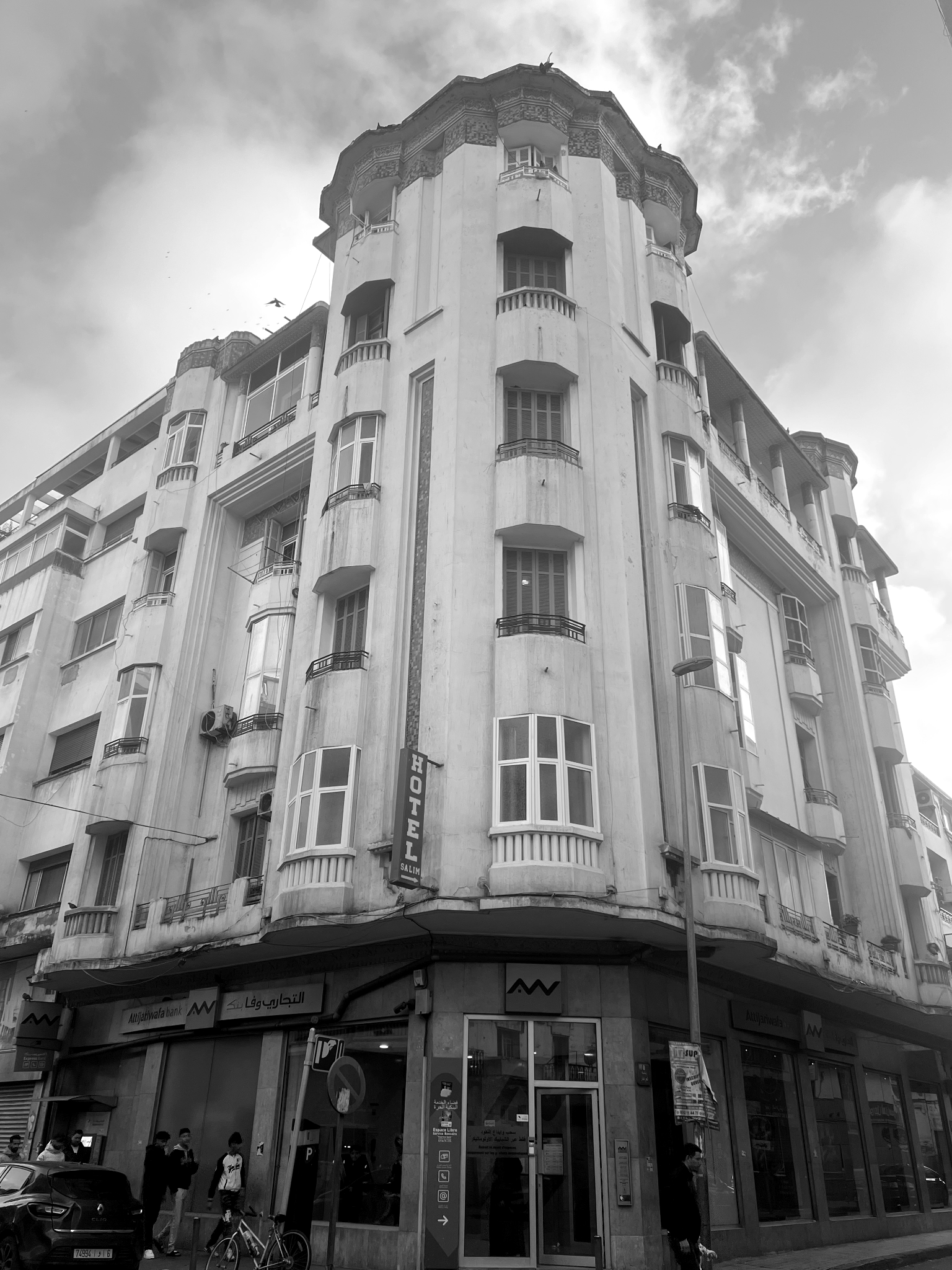A black and white photo of an old multi-story building with a hotel sign, large windows, small balconies, and some people walking on the sidewalk at the street level. Art residency casablanca