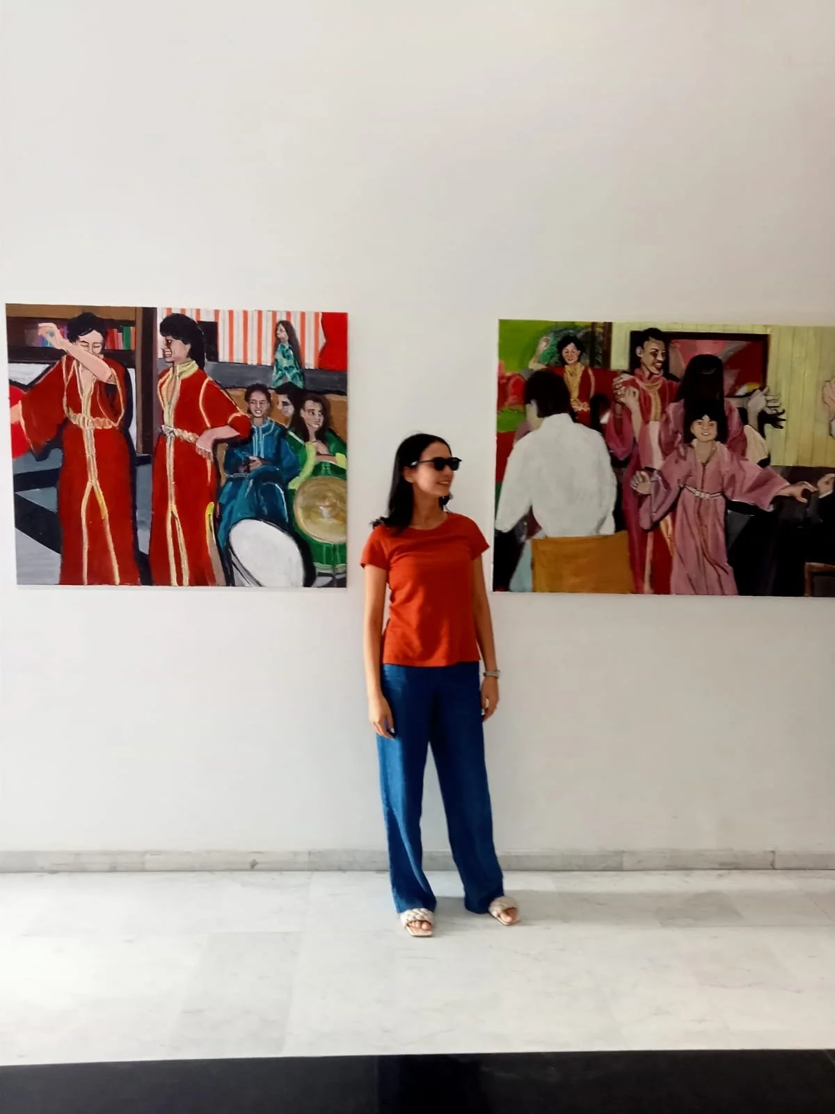A woman wearing sunglasses, an orange top, and blue jeans standing in an art gallery with colorful paintings of traditional Asian scenes on the wall behind her.