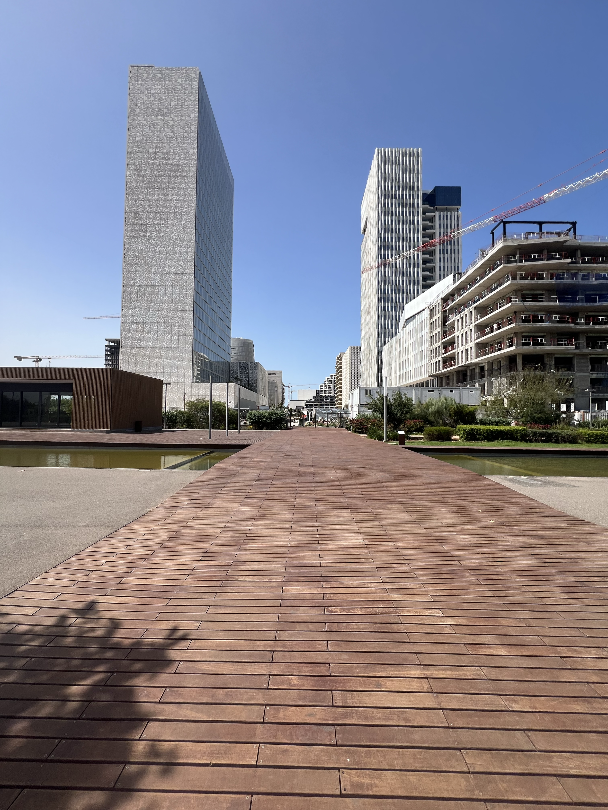 An urban park with a wooden pathway leading towards modern high-rise buildings under a clear blue sky.