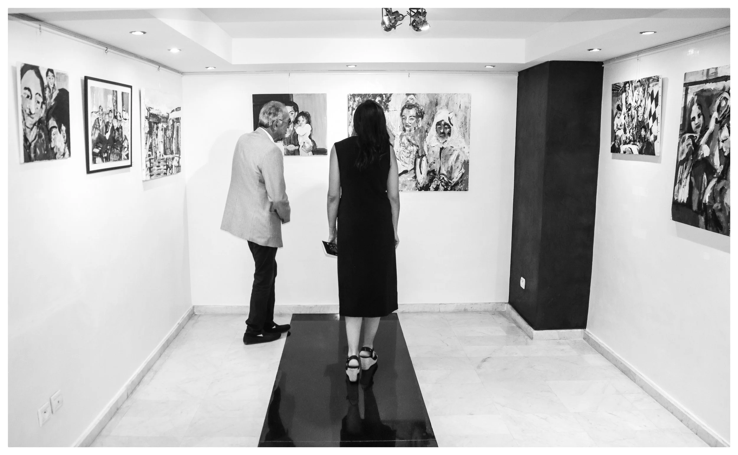 Two people viewing artwork in an art gallery with wall-mounted paintings, one man and one woman with a dark dress and high heels.