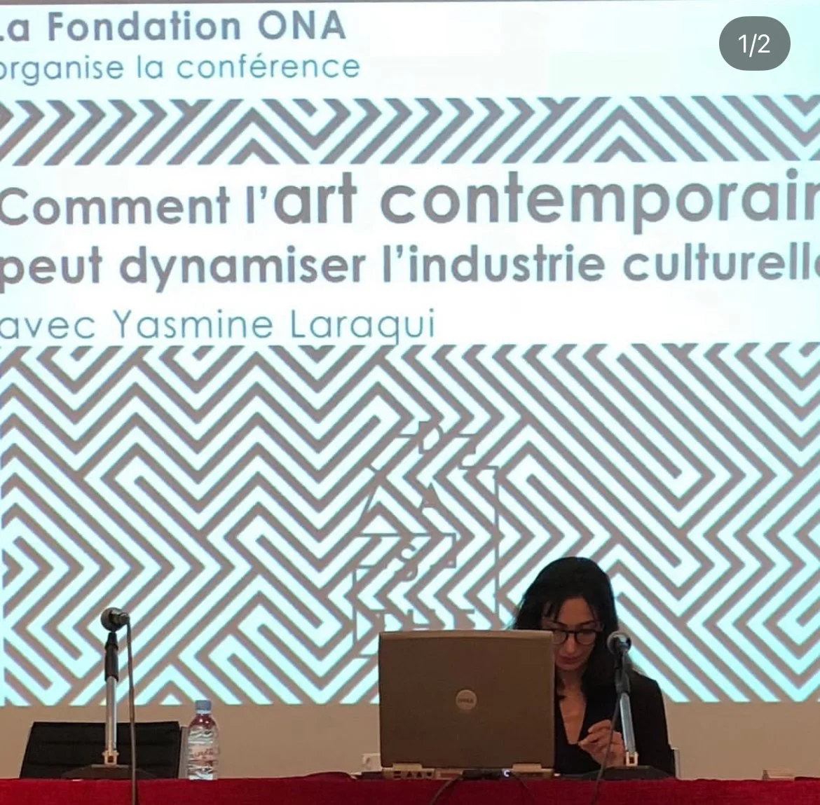 Conference presentation slide with French text about contemporary art and cultural industry, featuring geometric black and white patterns, a woman with glasses sitting at a table with a laptop and a microphone.