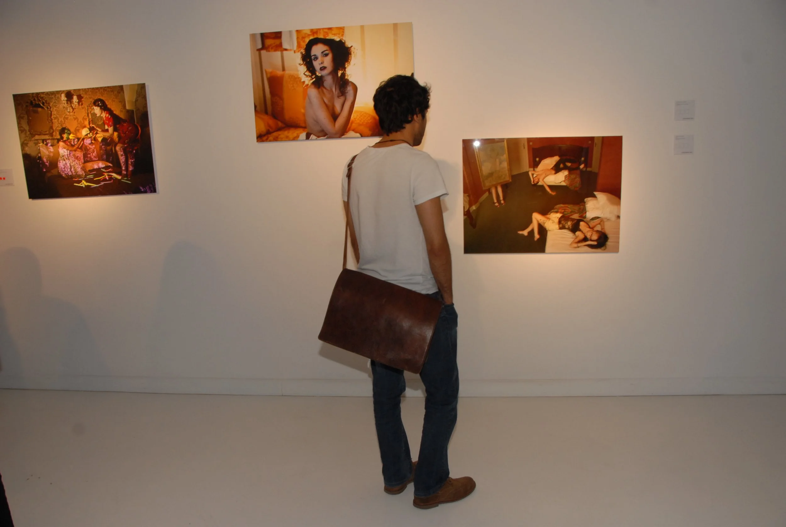 A man with curly hair, wearing a white T-shirt and jeans, stands in an art gallery looking at photographs of women. He carries a large brown bag over his shoulder.